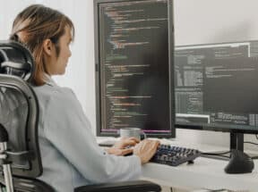 Young Asian woman software developers using computer to write code sitting at desk with multiple screens work remotely at home.